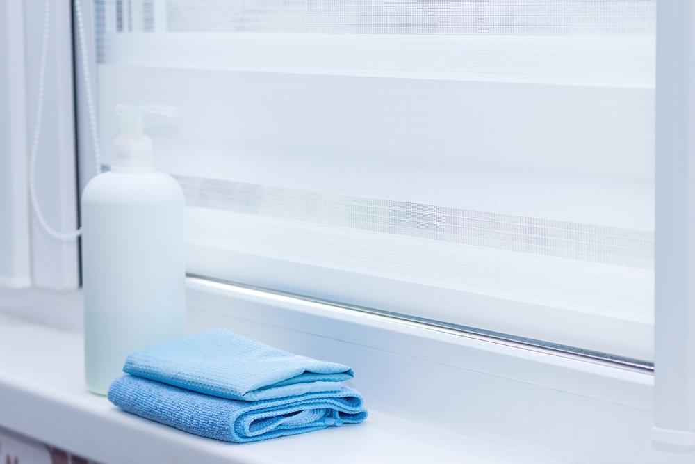 Cu ce se curăță jaluzelele orizontale din pânză: un pervaz alb de geam pe care sunt lavete albastre împăturite și un recipient cu detergent.