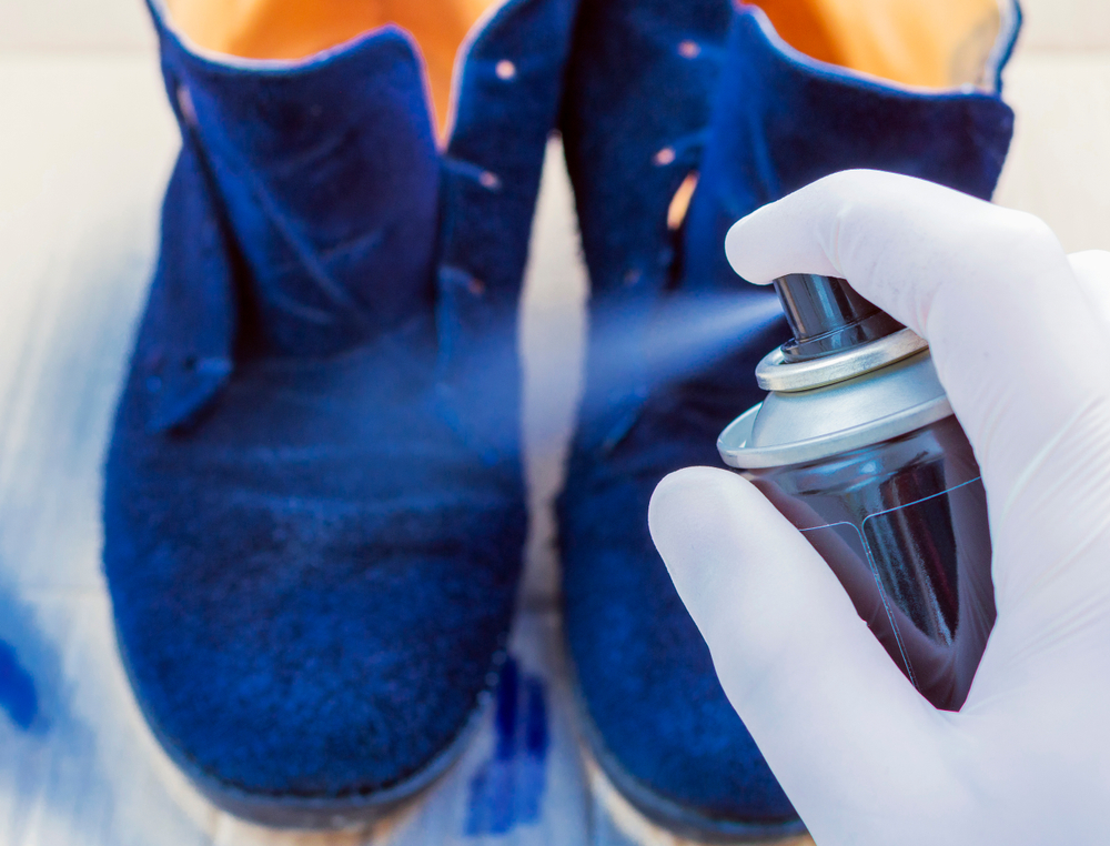 O mână cu mănuși albe pulverizează un spray de protecție peste ghete albastre din piele întoarsă.
