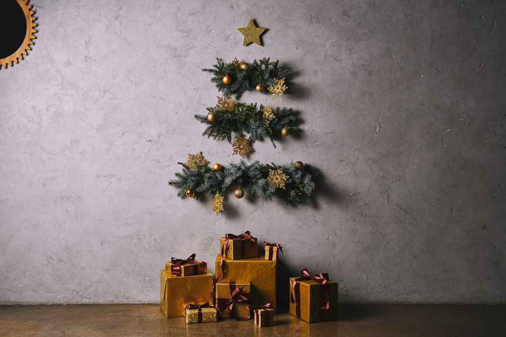 Un brad artificial pe perete realizat din ramuri de brad lipite de perete și decorate cu decorațiuni aurii și cadouri în cutii aurii la bază.