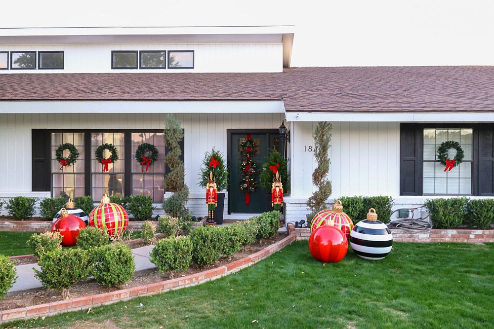 O grădină din fața casei decorată cu globuri supradimensionate. coronițe cu decorațiuni roșii și doi spărgători de nuci mari flanachează usa de la intrarea în casă. 
