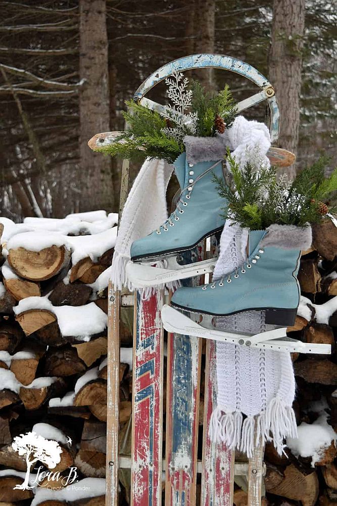 Idei decprațiuni Crăciun pentru grădină: O sanie vintage sprijinită de lemne înzăpezite, alături de patine de gheață bleu atârnate de sanrie, decorate cu ramuri de brad și un fular alb.