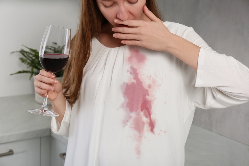 pata de vin pe haina alba făcută de femeie care bea un pahar de vin roșu