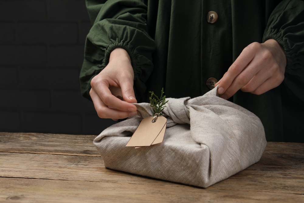 Idei de împachetat cadouri: Mâini de femeie ambalează un cadou în material textil cu o etichetă din carton maro deschis pe o masă din lemn natur, pe un fundal negru.